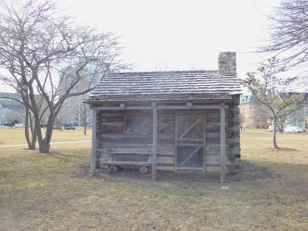 Exterior view of Solomon Juneau Replica Cabin in Juneau Park. The small log cabin has a covered porch and a log bench. A wooden entrance and window are on the front side. A stone chimney appears on the right side of the gable roof. The cabin sits on a lawn with trees around it. Buildings are visible in the far background.