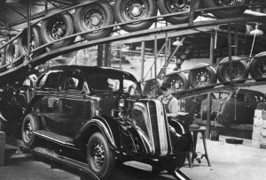 Long shot of a man working on a body of a vintage car inside the Nash Motors Assembly Line. Above the glossy-looking vehicle and behind the worker are rows of wheels held by a metal frame. Additional car parts are shown in blurry images in the background.