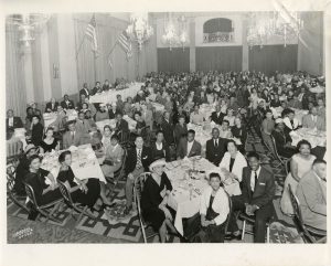 High-angle shot of the NAACP annual dinner showing the members of the Milwaukee branch making direct eye contact with the camera lens. The room is filled with people in formal attire sitting at dinner tables. Curtains hang on the wall in the background. Large antique ceiling lamps are glowing. Two American flags are placed on the wall on the left.