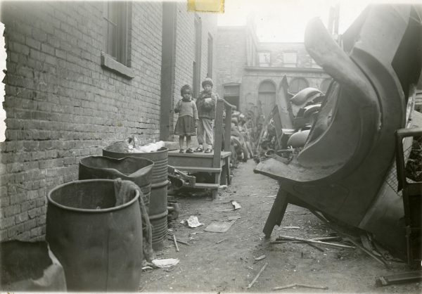Two small children stand on the rear porch of an apartment building once located on N. 8th Street. The yard is filled with scrap metal from junked cars.