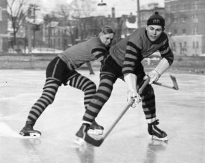 Sepia-colored full shot of John Cooper and Hank Kearns playing hockey in the horizontally striped uniforms. Each holds a hockey stick while gliding through an outdoor ice arena. Faraway buildings and leafless trees are visible in the background.