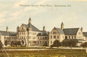 A painted postcard illustrates the grand building of the Milwaukee County Alms House grand building. The multiple-story building features gable and valley roofs. A green landscape spans the foreground.