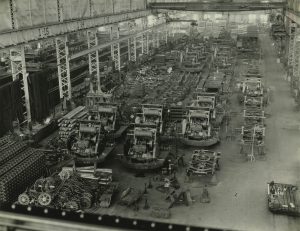 Sepia-colored high-angle shot of Pawling and Harnischfeger manufacturing plant interior. Rows of machines are placed in the large room. Gears and machinery equipment lies on the floor in the foreground. An aisle is visible on the right side. Steel infrastructure lines the left and right sides of the room.