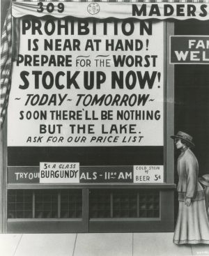 The Mader's Restaurant advertisement in grayscale urges people to stock up on liquor and beer. The painted ad illustrates a large poster installed on a sizeable window-like structure. The poster is filled with text written in big font. It reads, "Prohibition is near at hand! Prepare for the Worst, Stock Up Now! Today or Tomorrow, Soon There'll be Nothing but the Lake. Ask for Our Price List." Another announcement below the poster informs the price of burgundy and beers. Next to the poster is the image of a standing woman in a hat, a long-sleeve top, and a long skirt facing to the left.