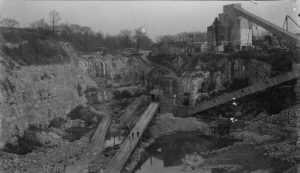 Elevated view of a deep basin in the area of the Certified Concrete quarry facility. Buildings and equipment appear in the basin and the area above the pit at the image's center and right. Two people stand on a road stretching down toward the pit's bottom. A body of water appears next to the road in the right foreground.