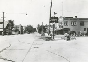 Grayscale long shot of the intersection of N. 35th Street and Silver Spring Avenue. A wide road stretches down the image's center. A hanging sign reading "pharmacy" and "Coca-Cola" appears on a building on the street's left side. A sidewalk sign inscribed Coca-Cola appears on the street's right side next to the "Wadhams Gasolines" sign. Commercial buildings line in the right background. Some cars traverse the street, and some are parked.