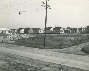 Grayscale long shot of a cluster of one-and-a-half-story houses under construction. Several ladders and workers are visible near the unfinished dwellings. An undeveloped space and a pile of rubble appear in the area closer to the camera lens. A utility pole and overhead wires are in the image's center. A road spans from left to right foreground.