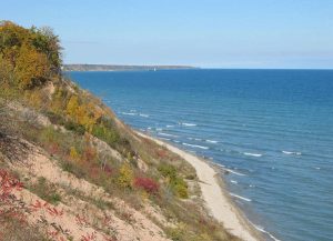 Panoramic view of a bluff in Lion's Den Gorge Nature Preserve set adjacent to the blue-colored Lake Michigan. A faraway view of Port Washington and the blue sky are visible in the background. A narrow beach or dirt path arcs between the bluff and the water's edge.