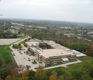 Bird's eye view of UW-Washington County Campus and its vicinity. The building complex and parking area are surrounded by green landscapes adorned with trees beginning to turn with fall colors. Other buildings appear in the far background.