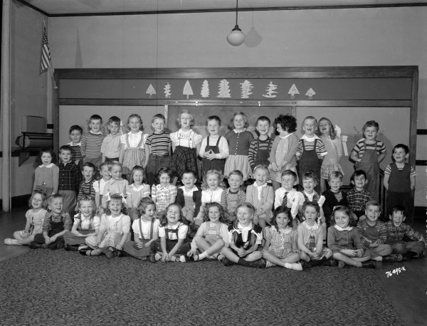 A grayscale group photo of dozens of Kindergarten students in three rows. Children in the first and second rows are sitting and those in the last row standing as they smile. An American flag is installed in the left corner of the room.