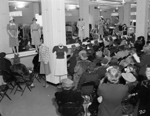 A grayscale long shot of women posing in dresses and hats on a catwalk stretching from left to right in Boston Store. Audiences gaze up at the models while sitting on rows of chairs in the foreground and background of this image.