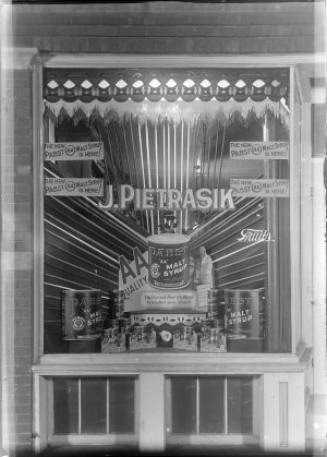 Grayscale photograph of a display window advertising the Pabst Malt Syrup product. Four rectangular papers that each read "The New Pabst 'AA' Malt Syrup is Here!" is placed on the window. Two are glued on the top left, and two are on the top right of the window. The papers flank a word written at the window's center that reads "J. Pietrasik." Beneath this are a variety of pictures and text promoting the Malt Syrup. Brick walls surround the window's left and upper sides.