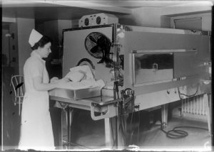 A nurse in uniform takes care of a patient whose body is inside a coffin-like cabinet called the iron lung at Milwaukee County General Hospital. The nurse stands on the left facing right with hands on the patient's head.