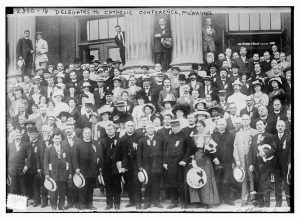 Grayscale group portrait of dozens of the American Federation of Catholic Societies members posing in formal clothes. They stand in rows on a building's front stairs. Five men in suits and ties stand near three giant columns in the back. The upper portion of the building's windows and its exterior walls are visible in the background.