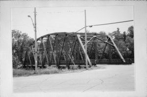 Grayscale long shot of the Newburg's steel truss bridge stretching in the distance at the image's center. The steel truss soars along and above the bridge's deck. Guard rails are installed on both sides of the bridge. A roadway spans from the foreground toward the deck. Two utility poles stand among weeds on the street's left side. Lush tall trees appear in the far background, across the bridge.