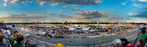 Panoramic view of the Milwaukee Mile during the NASCAR Camping World Truck Series. The image is taken from the grandstand, displaying the NASCAR tracks below in the distance. Above is a blue sky.