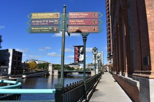 A panoramic view of Milwaukee's Riverwalk. A long metal fence with regularly spaced lighting poles separates the Milwaukee River on the left and a pedestrian area on the right. A directional signpost stands on top of the fence in the foreground.