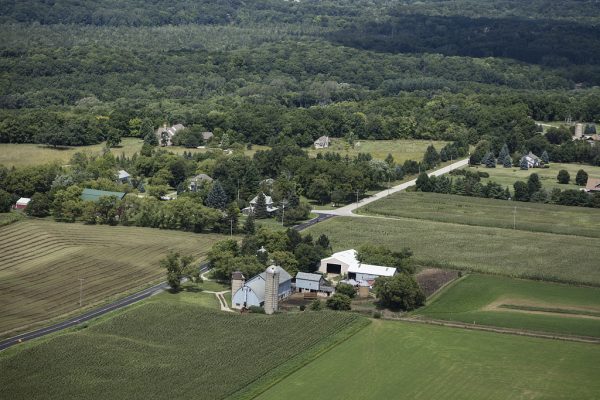 Bird's eye view of the a farm area in Lake Five community in the town of Lisbon. Green landscape fills most of the image. Sparse residential places are built among trees. A barn with a grain silo sits in the center foreground of the image.