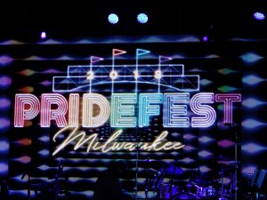 Long shot of a stage showing a composition of multicolor lights glowing in the dark. Neon lights in the top center form four flags and the number "2018." Beneath this is another neon light displaying the words "PrideFest" on the upper and "Milwaukee" on the bottom. Microphone stands and a drum set are slightly visible on the stage.