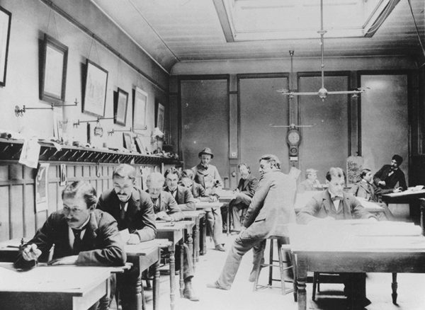 An office room full of the Ferry & Clas firm's workers in their suits and ties. Most of the men sit at their desks and concentrate on their tasks. At least three other people who look like the supervisors seem to oversee the employees' performance. One sits on a chair, and two stand near the workers.