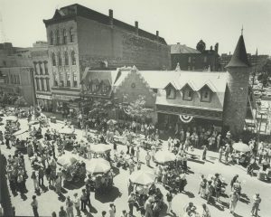 A high-angle shot of a block party held on a street in a grayscale color. People crowd the area. Some sit on rectangular tables. Some rest at round picnic tables that have umbrellas on them. Others stand here and there. Commercial buildings, including a furniture store and Mader's restaurant, line the background.