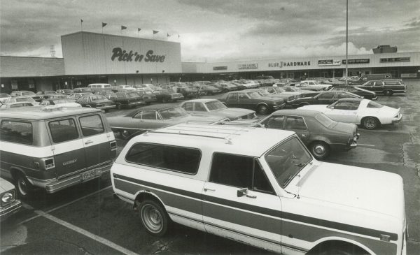 Wide shot of Greenfield's Spring Mall parking area in grayscale. Rows of cars park in the center, and the stores line the edge of the parking lot in the background of the photograph. Pick'n Save, Fish Galore, and Blue J Hardware storefront signs are among others visible in the mall's exterior wall.