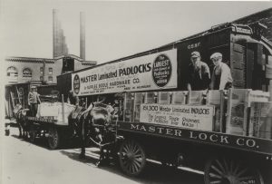 Side view of two horse-drawn vehicles carrying boxes containing Master Lock padlocks. A group of people load the packages onto a train with a large banner hanging on its side. Signs proclaim this "the largest single shipment of padlocks ever made." Inscribed on the wagons' bodies is "Master Lock Co." Four people standing on the wagons make direct eye contact with the camera lens.