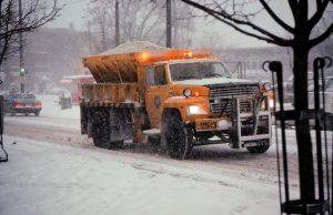 An orange-colored Milwaukee public work truck disperses salt on Downer Avenue on a snowy day. The truck faces to the right. The salt is placed in the salt spreader attached to the truck's rear. Snow falls and covers parts of the truck and the street. An area with thicker snow is visible in the foreground. Cars are visible in the foggy background.
