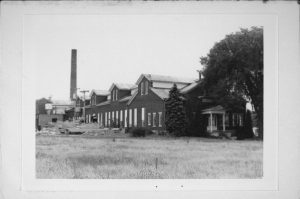 Long shot of the Morey Milk Condensery in grayscale. The entrance is on the right under the shade of a big, lush tree. The building extends to the left background. A tall chimney appears in the far left. A grass field spans the foreground.
