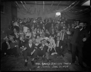 Grayscale group photo of dozens of people filling a room. They pose in different styles. Some hold a glass of drinks and bottles. One who holds an accordion sits next to a drinking man. One that plays concertina poses in the right background next to a door. Rows of ribbons decorate the ceiling and the light. Inscribed on the photo's bottom is "The Gang's Birthday Party at John's Jan., 20, 1934."