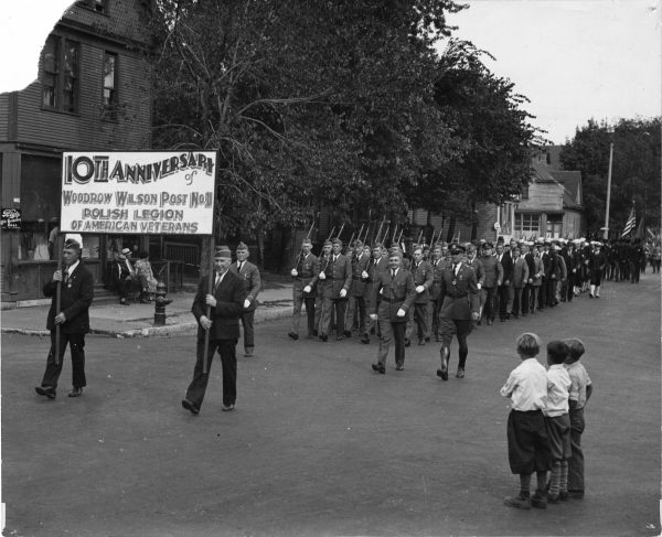 Grayscale long shot of rows of men in uniforms parading the street from right back toward the left. Two men walking in the front carry a banner that reads "10th Anniversary of Woodrow Wilson Post No. 11, Polish Legion of American Veterans." People marching in the front rows place a riffle on their right shoulders and hold the weapon's butt with their right hands. Three children stand in the right foreground as they watch the procession. Several buildings and trees are visible in the background. A small group of people sits in front of a building in the left background.