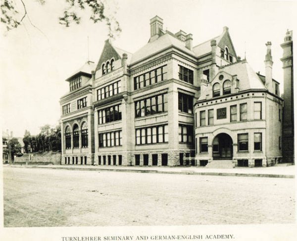 Sepia-colored exterior view of German-English Academy, a 3½-story building. The wing on the far left features three round-arched windows. Adjacent to the main building on the right is a 2½-story structure featuring a wide entrance and a the date "1892" inscribed on the second-floor exterior wall.