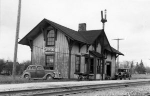 Grayscale long shot of the Sussex railroad depot behind a railroad track that spans the foreground. The simple wooden building features rectangular windows, a chimney, and a gable and valley roof. A baggage cart and two cars are placed around the depot.