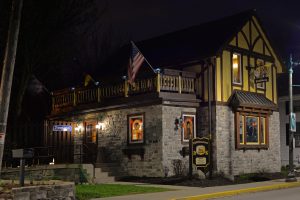 Exterior view of Von Rothenburg Bier Stube in Germantown at night. The two-story building features stone exterior walls composing its ground floor and a yellow-colored wall on the second. The first story has stained glass windows and a wooden entrance. A sign that reads "Biergarten" hangs near the front door. The balcony enclosed by balustrades has American and German flags on it.