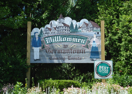 Outdoor sign made of wood is inscribed "Willkommen in Germantown." The words are set between a man and woman figure in traditional German clothing and under images of German-style buildings and fur trees. Next to it is a small board that reads "Best Places to Lives, Money Magazine's 2007 Great American Towns."