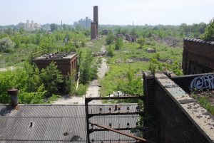 High-angle shot of Solvay Coke ruins scattered on a wide abandoned ground among unkempt plants. Upper parts of a building with its rusty components appears in the foreground.