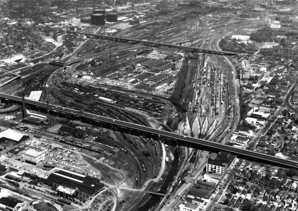 Aerial shot of Menomonee Valley in grayscale tone. The image shows a busy industrial area. Visible from afar are two long highways, crossing over two large sets of railroad tracks partially occupied by trains. Different kinds of industrial infrastructure appear in the foreground and background of the photograph. A portion of a residential area is visible on the bottom right of this photograph.