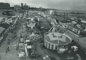 Garyscale high-angle shot of the 1971 Summerfest's midway showing various attractions in the image's center. The crowd walk on streets that flank the attractions arena in the center. Milwaukee's buildings are visible in the far left and center background. Lake Michigan appears on the far right.