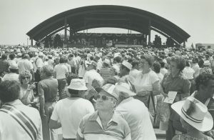 Grayscale long shot of a large crowd standing in summer clothes in front of Festa Italiana outdoor stage. The stage in the far background is covered by an arched roof.