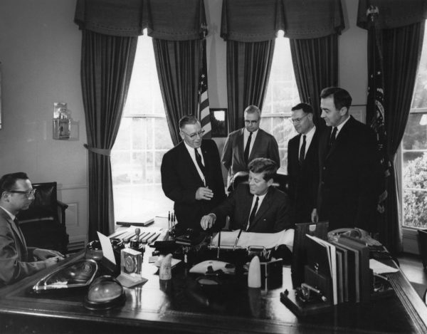 Grayscale photograph of four men in formal attire standing behind President John F. Kennedy in the Oval Office. Representative Henry Reuss in glasses stands second from the right. President Kennedy smiles as he signs the document on the desk. A man sits on the far left with his body facing the President. A pole with an American flag appears around the center back. Light emanates through the three curtained windows in the background.