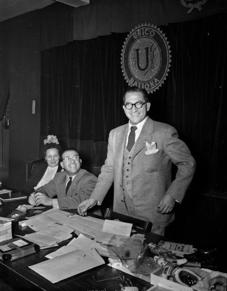 Grayscale portrait of William Calvano smiling in a suit and tie. He stands beside Joseph Bruno and Dorothy Matranga, who sit next to a long desk full of documents. Hanging behind them is a circular UNICO logo.