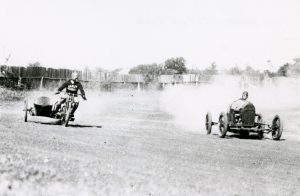 Grayscale wide shot of the rider of a vintage motorcycle on the left racing with an antique car on the right. Both leave behind a cloud of dust . The motorbike rider wears a Harley Davidson jacket. His vehicle features a sidecar. Both drive through the corner of the dirt-surfaced track. A fence lines the circuit in the far background.