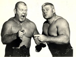 A photo montage showcases Dick "The Bruiser" Afflis and Reggie "The Crusher" Liswoski. This photo of the two wrestlers is made as if they are standing next to each other. They appear in wrestling belts, glaring while pointing in different directions with their forefingers.