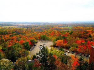 Elevated shot of a scenic view taken from the Holy Hill National Shrine building. Trees in fall colors can be seen as far as the eye can see. A parking area filled with several cars is visible in the center-to-right of the image.