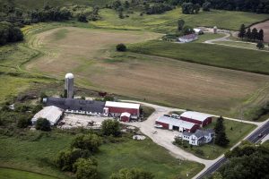 Bird's eye view of a farm in Washington County. Several buildings and a silo surrounded by a green landscape appear in the foreground. Extensive arable land and green areas are visible in the background. Other low buildings and a silo can be seen in the right back.