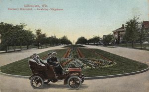 Postcard showcasing a panoramic view of Newberry Boulevard. A vintage automobile carrying passengers appears in the foreground next to a wide green lawn adorned with landscaping flowers set in the middle of the boulevard. Tall trees grow along the road verge in the left and right side of the street.