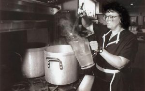 Medium shot of Barbara Franke ladling boiled water from a stock pot into a pitcher inside Miss Katie's Diner's kitchen. She wears glasses standing next to a stove with two stock pots on it. Steam rises from the pitcher.