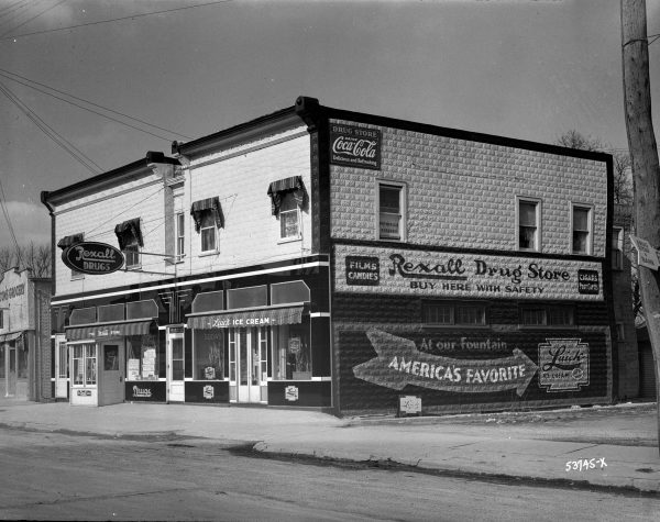 Grayscale long shot of a two-story building facing slightly to the left. The image shows two sides of the structure. The one on the left is the facade. It features three entrances on the ground floor and several identical rectangular windows on the upper floor. The door on the left belongs to Rexall Drug Store. A smaller door in the center has a sign with a physician's name. The right entrance is for the Luick ice cream shop. The building's other sides have a mural advertising the drug store and ice cream parlour on the ground floor. A sign promoting Coca-Cola is installed on the second floor's top left corner. A street stretches from left to right in the foreground. An adjacent building and overhead utility wires are visible on the far left.
