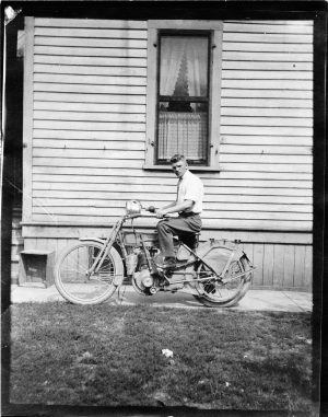 A side view of a man sitting on an early 20th-century motorcycle with an exterior wall of a structure as the background. He poses next to the building's window, on a pathway that separates the building with a lawn in the foreground. While his body facing to the left, his face gazes at the camera lens.
