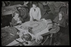 Grayscale high-angle shot of four women working around a long table full of crochet rugs. The faces of three women are visible in this image. The women on the left and right are sitting, and the one in the middle is standing. The fourth woman's back is partially visible in the image's foreground. Other tables with people working on rugs appear in the background.